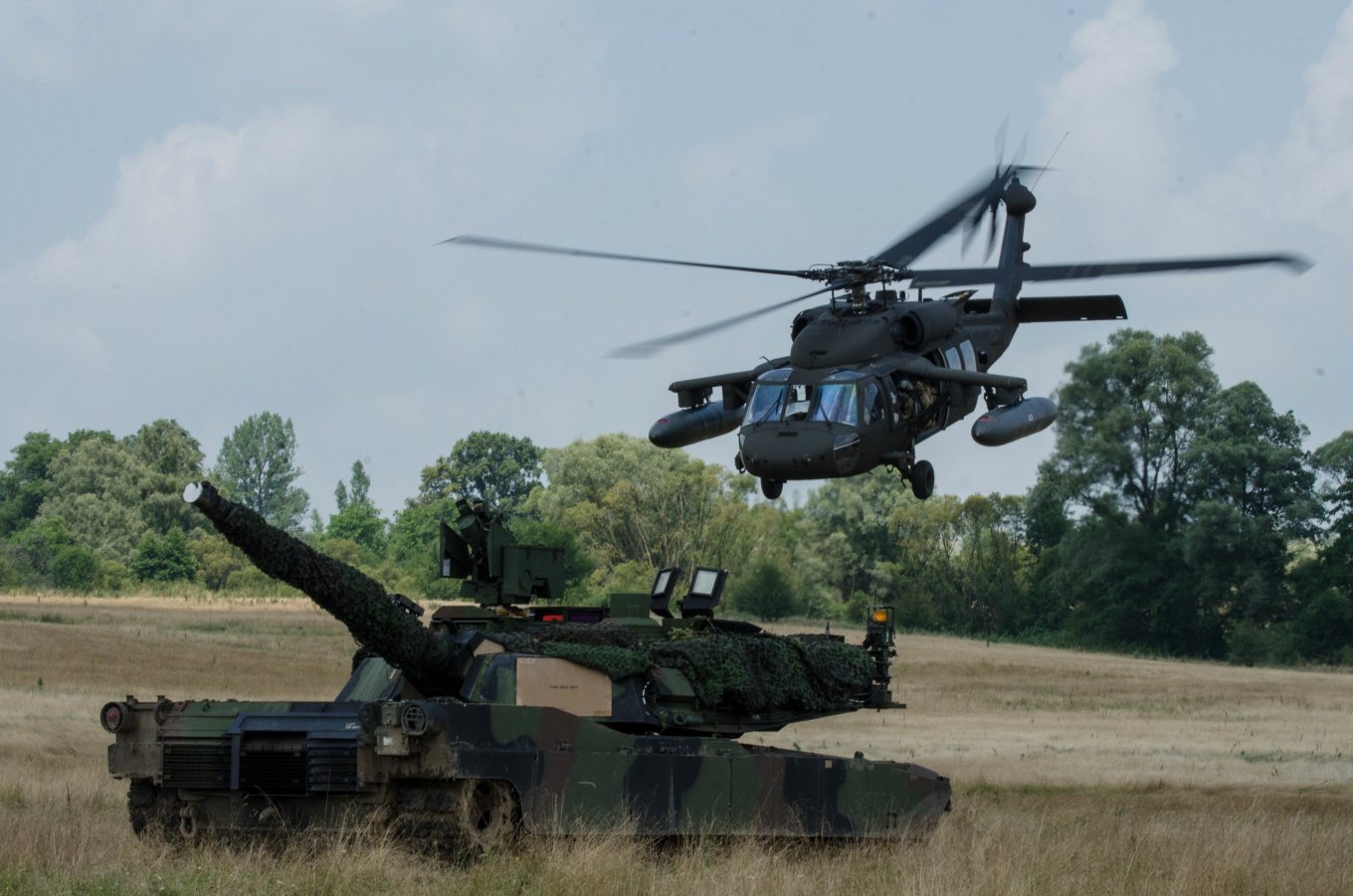 A UH-60 Blackhawk helicopter flies over a M1A2 Abrams main battle tank